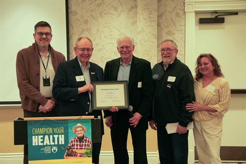 Male award recipients hold award certificate standing in the middle of found other individuals behind a podium