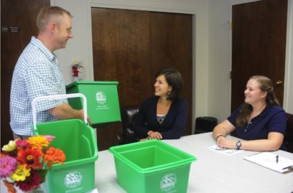 Person handing out recycling bins to people sitting at a conference table.
