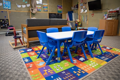 A white table surrounded by six blue chairs on a multi-colored carpet in a daycare-type setting.